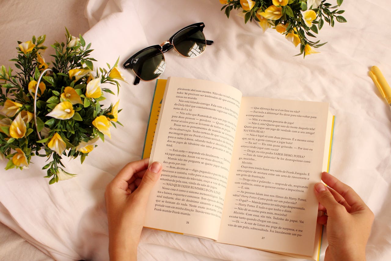 Home Hands holding a book surrounded by yellow flowers and sunglasses on a sunny day.