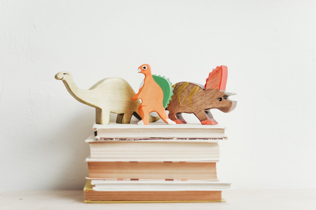 Home Colorful wooden dinosaurs on top of a stack of hardcover books on a light background.